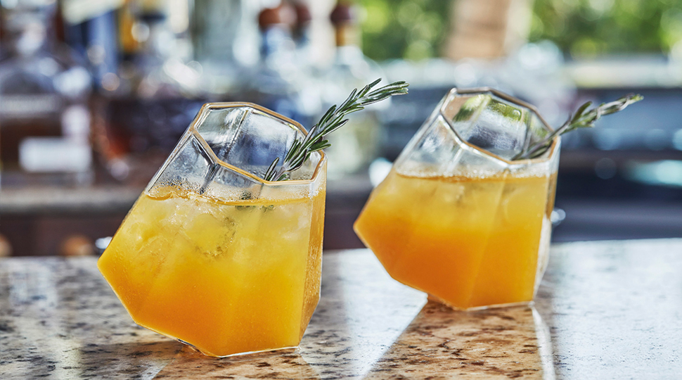 Two octagonal cocktail glasses each filled with a orange-coloured beverage and garnished with a spring of greenery