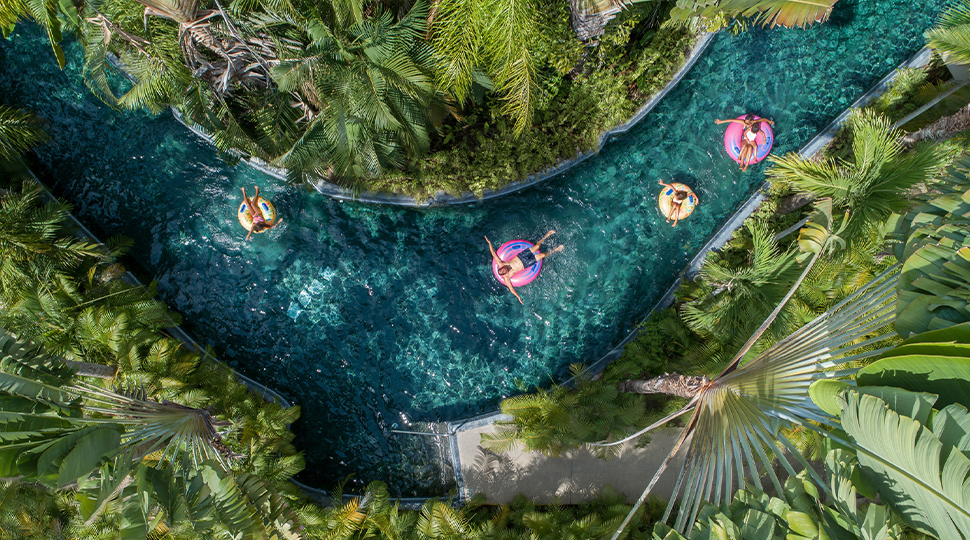 Aerial view of three four people floating in pink and yellow inner tubes along a lazy river surrounded by greenery