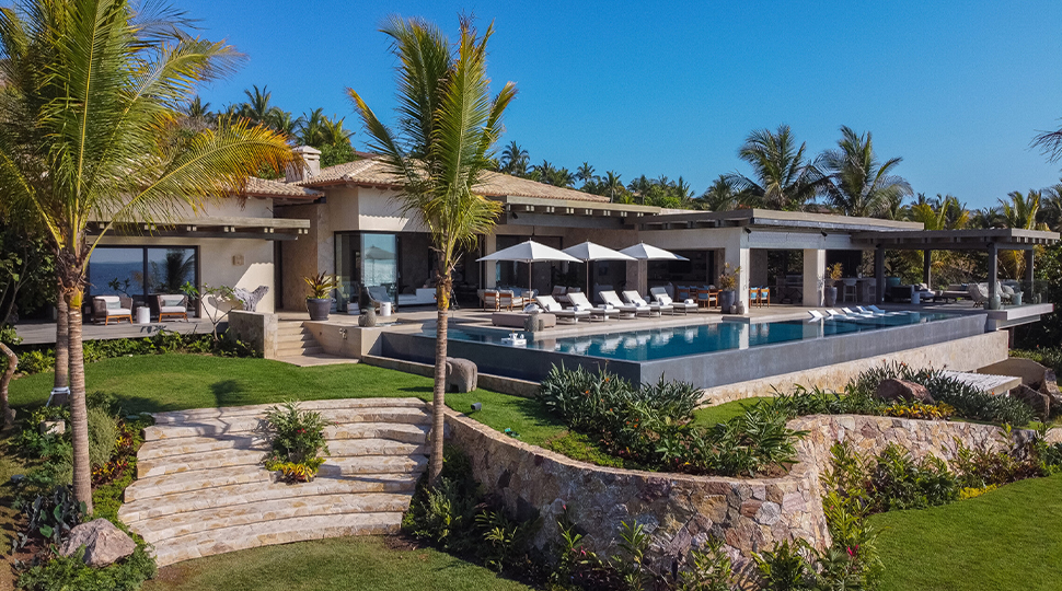 Exterior of a private villa with an infinity pool, white lounge chair and umbrellas and a small stone staircase leading down to a lawn