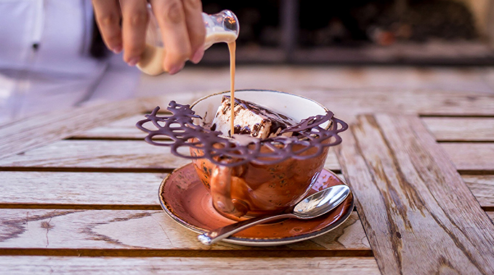 Hand pouring liquid into a brown mug of hot chocolate topped with a large piece of chocolate lattice and large marshmallow