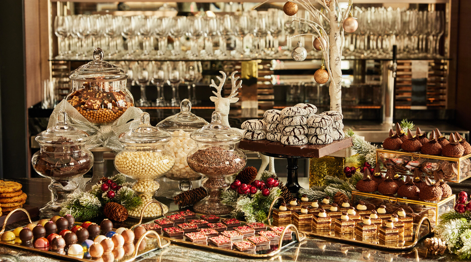 Large display of six trays of holiday desserts including a variety of pastries and cookies as well as five glass jars filled with candy and other sweets