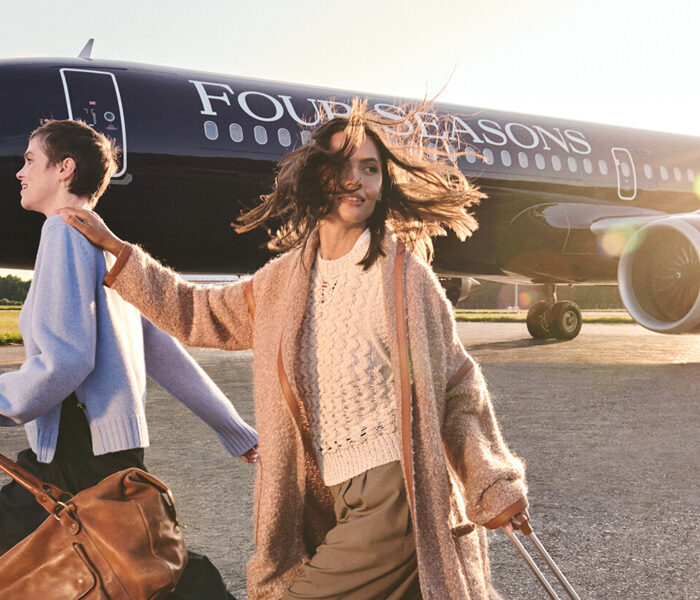 Two people wearing light-coloured sweaters and pulling rolling suitcases walk on the tarmac with a black Four Seasons branded private plane behind them