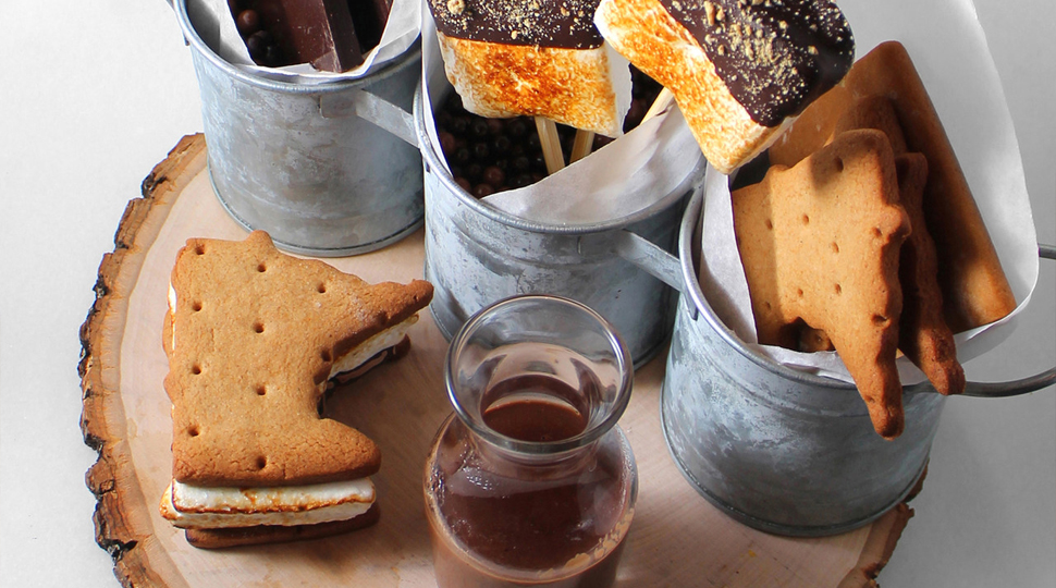 A wooden tray holds two Minnesota-shaped s'mores, three aluminum mugs, two crispy marshmallows on sticks, chocolate bars and a small glass carafe of chocolate sauce