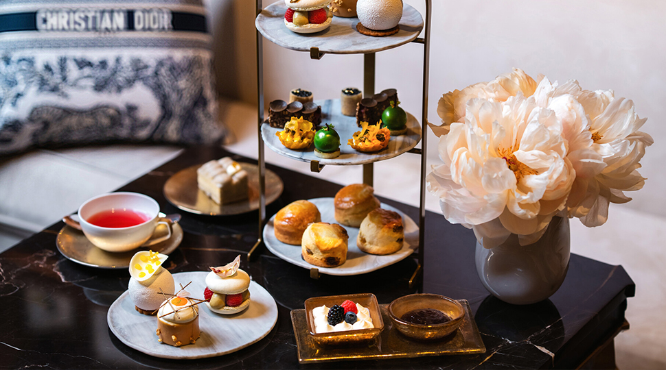 Three-tiered marble tray filled with assorted pastries, a cup of tea, three small plates of pastries and a vase of pink flowers all sit on a small black table