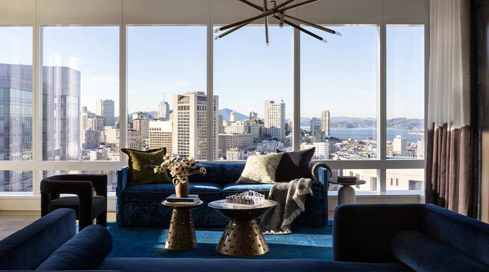 Blue velvet sofas atop a blue rug with two small copper coffee tables are in the centre of a living room with floor-to-ceiling windows in the background overlooking a city skyline