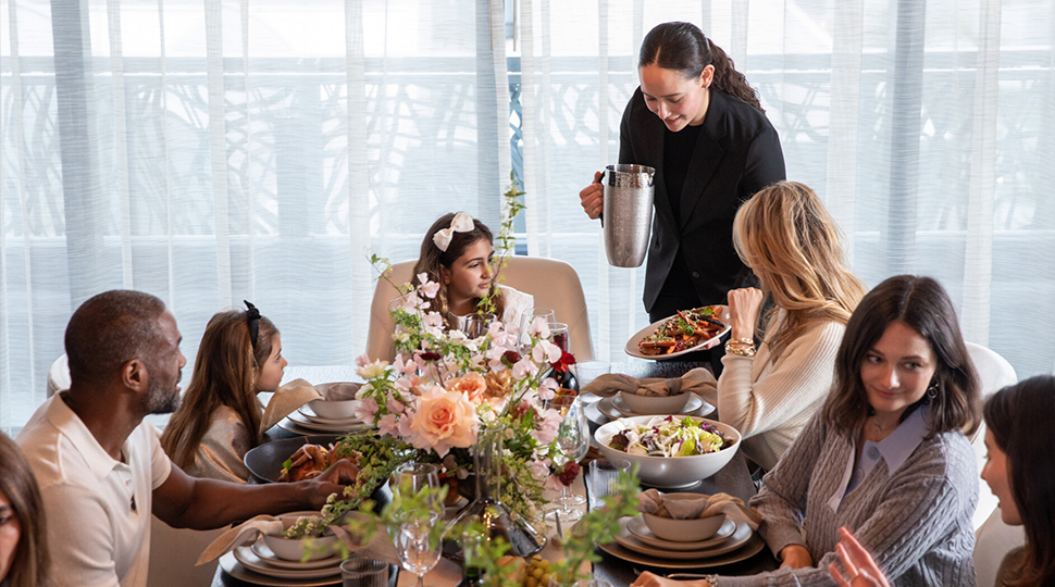 Six people sit at a table with a large peach and pink floral centrepiece as a server brings a plate of food to the table