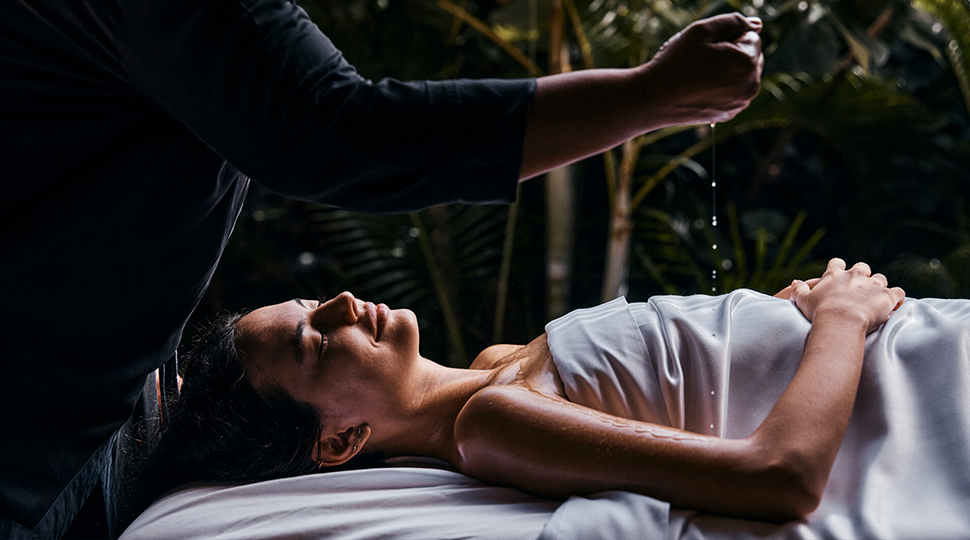 Person lays on their back under a silver satin sheet with their eyes closed as a pair of hands pours liquid over their arm