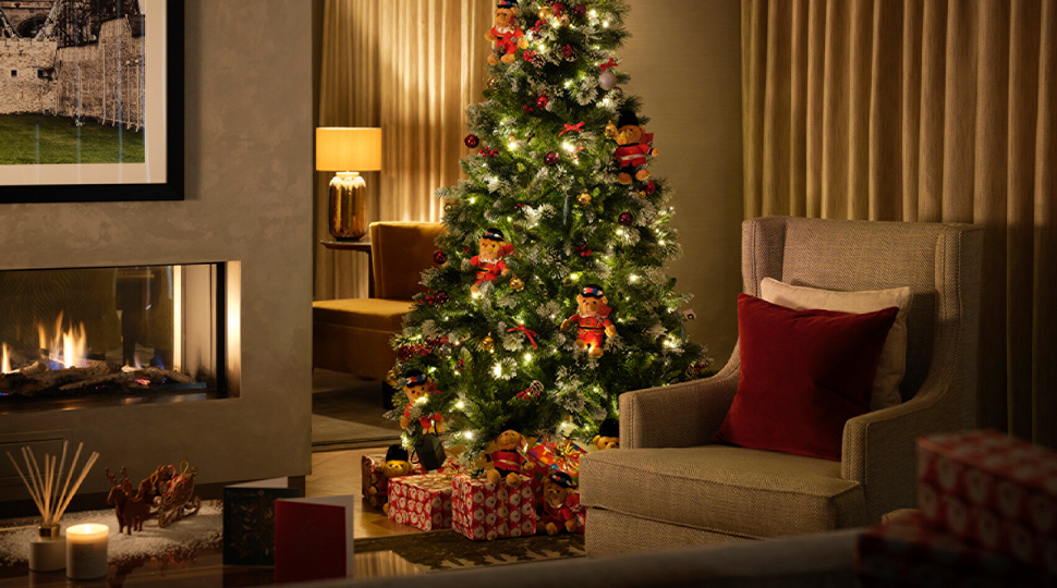 Christmas tree decorated with yellow twinkle lights, red and silver ornaments, and six stuffed bears wearing red uniforms, all set next to an grey arm chair and a fireplace